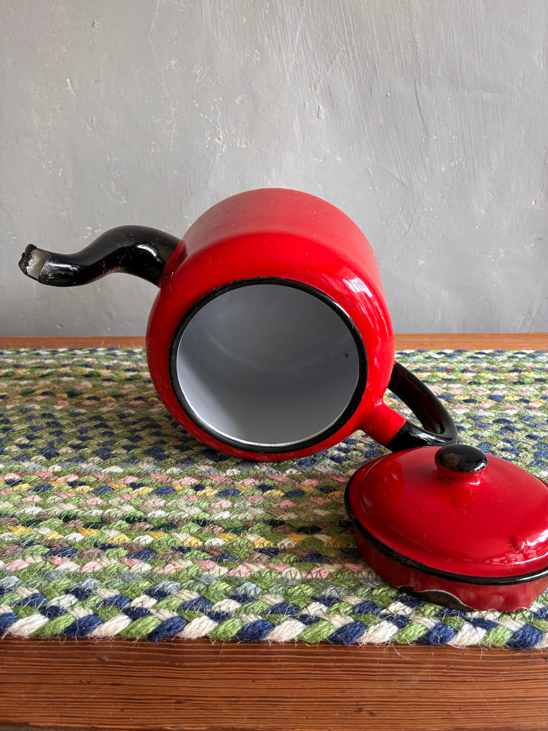 Vintage red enamel teapot with black handle and spout, showing white interior, resting on woven mat.