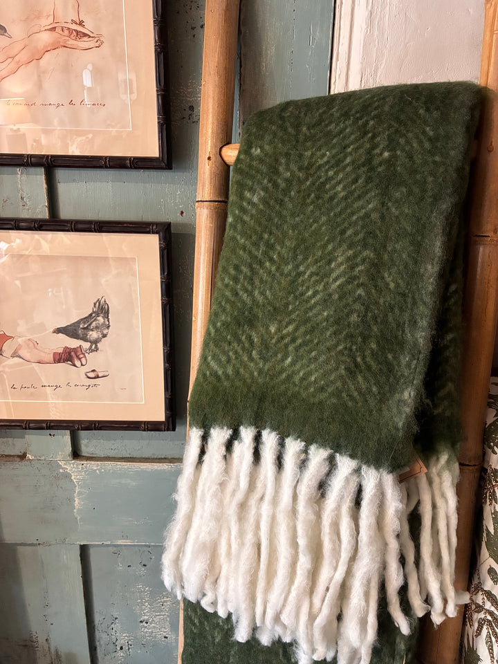 Green woven blanket with white fringe draped over a wooden rack against a rustic wall with framed pictures.