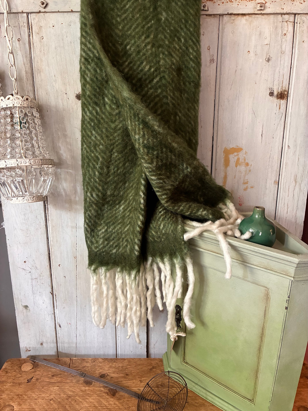 olive green throw with white tassels hanging against a wooden wall with a green corner cupboard in front
